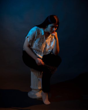 A girl stands on a miniature Greek column illuminated by multicolored light