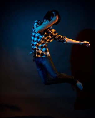 A young man in a plaid shirt jumps on a dark background illuminated by blue and yellow light