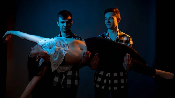 Two guys in a plaid shirt are holding a girl on a dark background in a studio with multicolored light — Photo