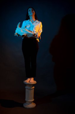 A girl stands on a Greek column with a bull skull in her hands illuminated by blue and yellow light