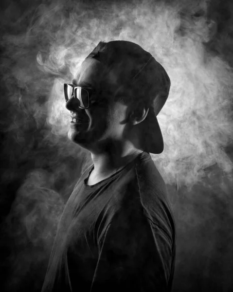 Ασπρόμαυρο σκιαγραφικό πορτραίτο ενός νεαρού άνδρα με γυαλιά και καπέλο στο φόντο του φωτισμένου καπνού — Φωτογραφία Αρχείου