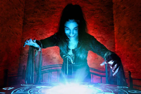 Die Hexe zaubert auf einem geheimnisvollen Tisch im steinernen Keller eines geheimnisvollen Hauses, das von blauem und rotem Licht erleuchtet wird Stockbild