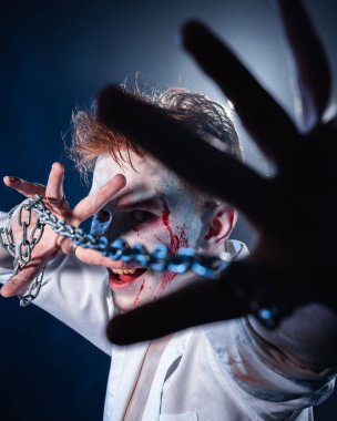 Makyajlı ve yüzünde kan olan bir manyak, saldırganlık zinciri olan bir adam fenerdeki duman ve parıltının arka planına doğru kameraya bakıyor.