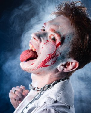 Yüzünde saldırganlıkla makyaj yapan bir psikopat kameraya mavi dumanın arka planına bakıyor.