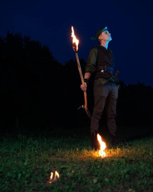 Peter Pan gibi giyinmiş bir adam elinde yanan bir meşaleyle gece gökyüzüne ve karanlık ormana karşı duruyor.