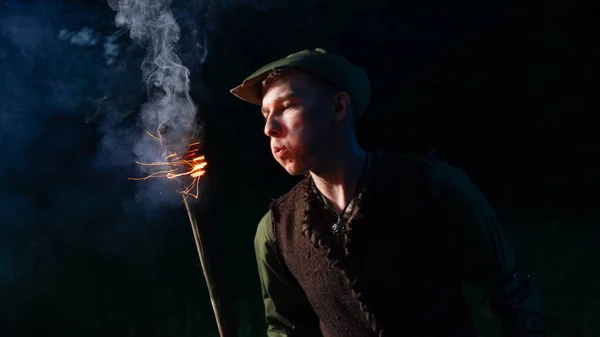 Un tipo vestido como Peter Pan está de pie con una antorcha ardiente contra el cielo nocturno y el bosque oscuro — Foto de Stock