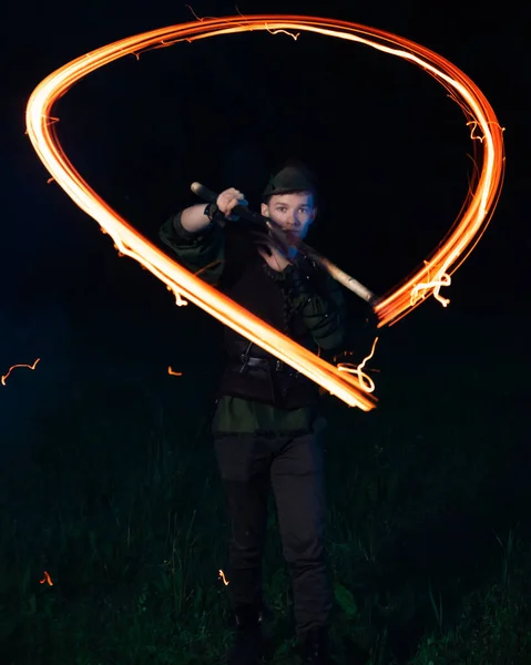 Um tipo com um fato de Peter Pan torce uma tocha em chamas nas mãos, o que deixa um rasto ardente, noite contra a floresta escura. — Fotografia de Stock