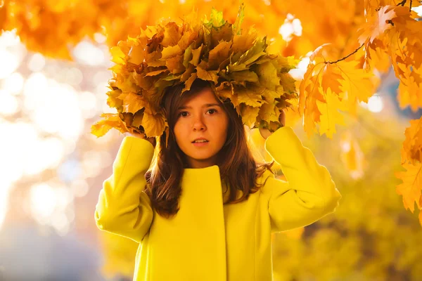 Chica con un abrigo amarillo llevaba una corona de hojas de otoño brillantemente caídas en el bosque — Foto de Stock