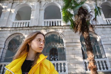 Sarı ceketli sarışının portresi eski bir binanın arka planında ve palmiye ağacında. Seyahat ve şehir turizmi.