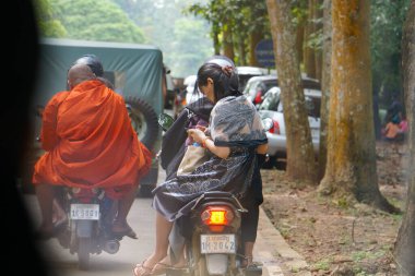 Siem Reap, Kamboçya - 6 Kasım 2025 - Geleneksel elbiseli genç bir Khmer kadın, şoförün arkasında motosiklet sürerken telefonunu kullanıyor, ve Angkor Tapınağı yolunda başka bir bisikletçiyle birlikte Budist bir keşiş