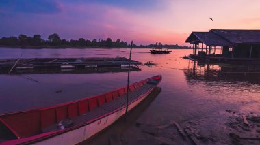 Ahşap bir yolcu gemisi gün batımında Mahakam Nehri 'nde süzülüyor. Önünde demirli bir kano ve renkli gökyüzüne karşı nehir kenarında bir ev silueti var..