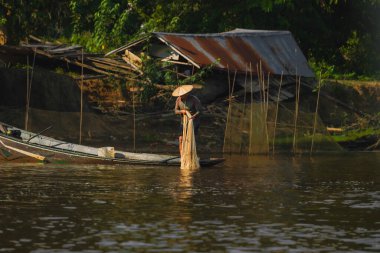 Kutai Kartanegara, Endonezya - 10 Ekim 2025. Geleneksel konik şapka takan yerel bir balıkçı sıcak bir sabah ışığında sakin bir nehir kenarındaki ahşap bir tekneden ağlarını atmaya hazırlanıyor..