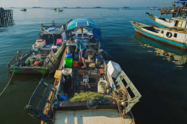 Balikpapan, Endonezya - 8 Kasım 2025. Yükselmiş bir manzara, sakin denizdeki diğer birkaçının yanında demirlemiş olan geleneksel teknesinin dağınık ahşap güvertesinde konteynerlerin ve balıkçı malzemelerinin arasında çalışan bir balıkçıyı yakalıyor..