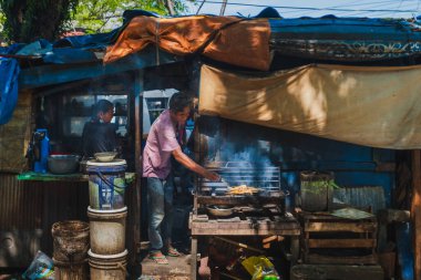 Balikpapan, Endonezya - 10 Aralık 2025. Pembe gömlekli bir erkek sokak yemeği satıcısı, derme çatma mavi ve turuncu muşamba bir barınağın altında açık bir kömür ocağının üzerinde balık ızgara yapıyor. Yıpranmış ahırın arka planında çalışan bir kadın.