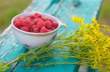 Güzel bir fincanda taze toplanmış ahududu ve bir buket yabani çiçek. 