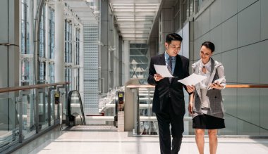 Ofiste yürüyen ve kağıt okuyan iki gülümseyen meslektaş. Modern ofis binasında elinde kağıtlar ve dijital tabletle yürüyen mutlu iş arkadaşları.