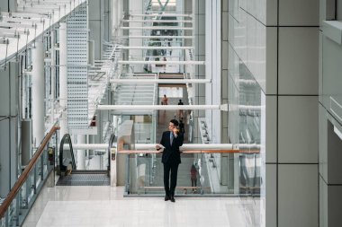 İş ortağıyla akıllı telefondan konuşan ve modern ofis binasında tablet tutan bir iş adamı mavi takım elbiseli şık bir iş adamının gelecekteki ofis binasında çalışmak için cep telefonu ve dijital tablet kullandığını görüyor.