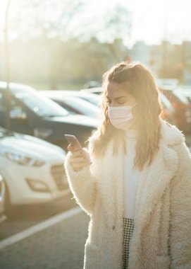 Koruyucu yüz maskesi takan ve otoparkta cep telefonuna mesaj yazan genç bir kadın.