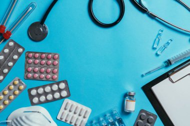 Mavi arka planda tıbbi ekipmanlar var. Düz steteskop, bez, hap, şırınga, aşı, ampul, dosya ve yüz maskesi.