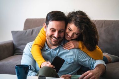 Birlikte telefona bakan mutlu çift. Gülümseyen kız arkadaşı mutlu erkek arkadaşını kucaklarken akıllı telefon kullanıyor ve gülüyor.