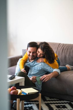 Mutlu çift evde akıllı telefon kullanıyor. Gülümseyen kız arkadaşı mutlu erkek arkadaşının arkasından sarılıp cep telefonu kullanıyor ve evde birlikte gülüyor.