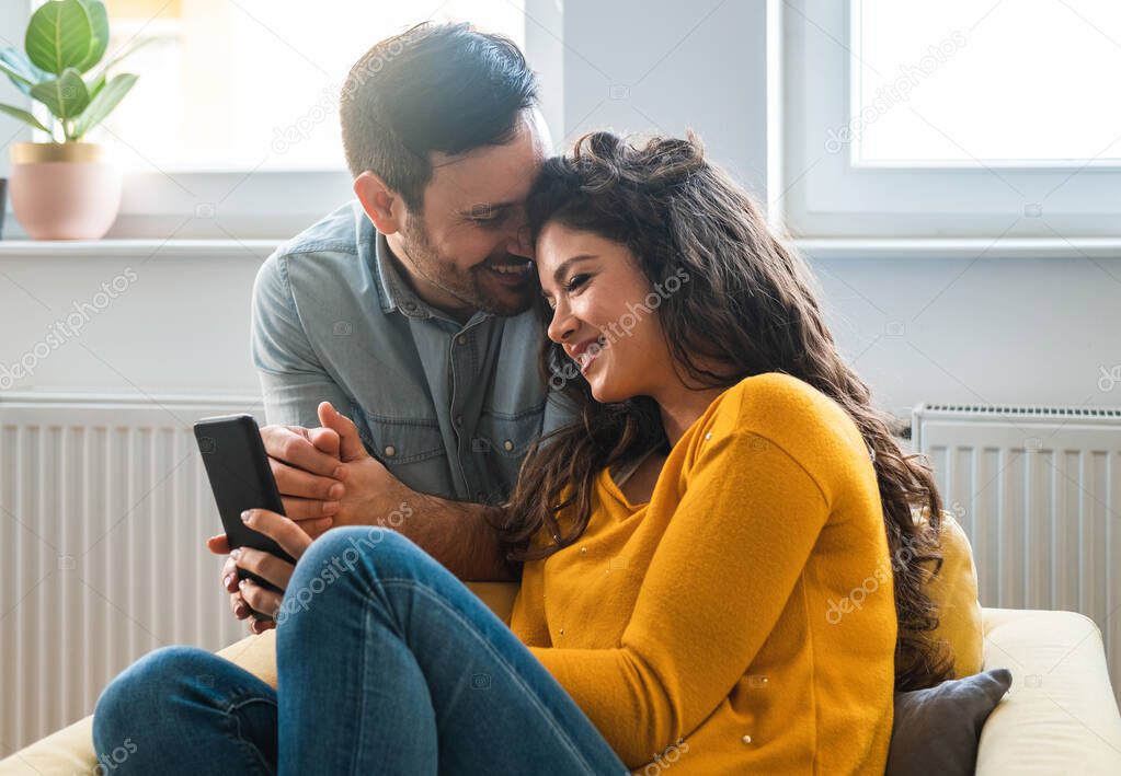 Loving Couple looking in smartphone together at home. Happy couple looking in mobile phone and enjoying in modern living room