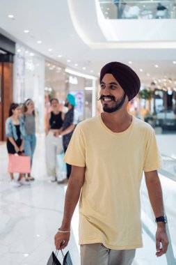 Yakışıklı Hintli adam alışverişi seviyor. Mutlu Hintli Sih adam alışveriş merkezinde yürürken sarık takıyor ve alışveriş torbaları tutuyor.