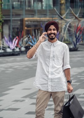Cep telefonuyla konuşan ve dışarıda alışveriş torbası tutan yakışıklı bir Hintli. Alışveriş merkezinin önünde durup akıllı telefonuyla konuşan gülümseyen adam.
