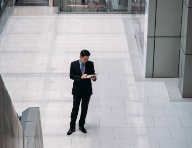 Modern ofis salonunda dijital tablet kullanan yakışıklı işadamı. Asyalı iş adamının tablet üzerinde ayakta durup mesaj ya da e-posta yazarken görüntüsü