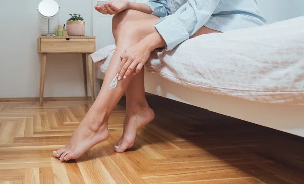 在卧室里 女人在腿上涂奶油 把无法辨认的年轻女人坐在床上 给她的腿和身体涂上润肤乳液的镜头剪下来 — 图库照片
