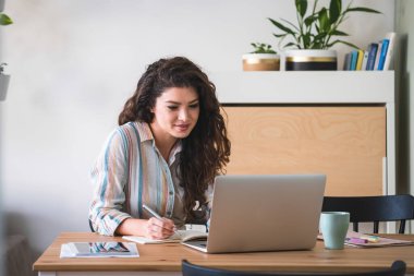 Dizüstü bilgisayarında video görüşmesi yapan mutlu bir kadın. Gülümseyen kadın iş arkadaşlarıyla video konferansı yapıyor ve notlar yazıyor. Kilitleme konsepti sırasında evden çalışmak