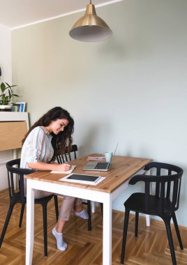 Evden çalışan güzel bir iş kadını. Sırıtkan iş kadını dizüstü bilgisayar ve tabletle masada oturuyor ve notlar yazıyor. Arkadaşlarla ya da iş arkadaşlarıyla çevrimiçi ders ya da video çağrısı almak