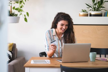 Evde elinde kredi kartı tutan ve dizüstü bilgisayar kullanan gülümseyen bir kadın. Masasında kredi kartıyla oturup online alışverişe para harcayan heyecanlı bir kadın.