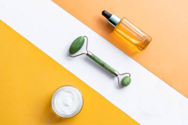 Yüz kremi, yeşim taşı silindiri ve geometrik arka planda yüz serumu. Kadınlar için yaşlanma karşıtı cilt bakımı ürünü. C vitamini, hyaluronik asit veya retinol serumu, Q 10 yüz nemlendiricisi ve çok renkli arka planda yeşim yuvarlayıcı.