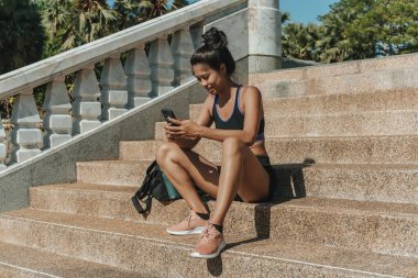 Antrenman molası vermek: Aktivite kıyafetli gülümseyen kadın merdivenlerde oturuyor ve antremandan sonra performansı izlemek için cep telefonu fitness uygulamasını kullanıyor. Sportif Asyalı kız, açık hava eğitiminden sonra gelişimi kontrol etmek için akıllı telefon kullanıyor.