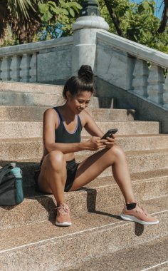 Antrenman molası vermek: Aktivite kıyafetli gülümseyen kadın merdivenlerde oturuyor ve antremandan sonra performansı izlemek için cep telefonu fitness uygulamasını kullanıyor. Sportif Asyalı kız eğitimden sonra ilerleyişi kontrol etmek için akıllı telefonu kullanıyor, internetten arkadaşını arıyor.