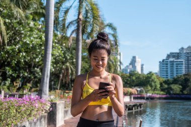 Antrenman molası almak: Aktif giyimde ayakta duran ve antremandan sonra performansı izlemek için cep telefonu fitness uygulaması kullanan gülümseyen bir kadın. Antrenmandan sonra ilerleyişi kontrol etmek, mesaj yazmak veya çevrimiçi arkadaşını aramak için akıllı telefon kullanan Asyalı kız