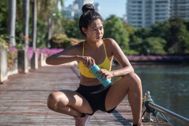 Genç bayan koşucu güneşli bir sabahta antrenmandan sonra dinleniyor. Kadın koşucu elinde su şişesi, koşuya ara veriyor, şehirden uzağa bakıyor.