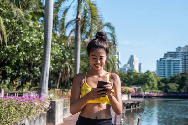 Antrenman molası almak: Aktif giyimde ayakta duran ve antremandan sonra performansı izlemek için cep telefonu fitness uygulaması kullanan gülümseyen bir kadın. Antrenmandan sonra ilerleyişi kontrol etmek, mesaj yazmak veya çevrimiçi arkadaşını aramak için akıllı telefon kullanan Asyalı kız