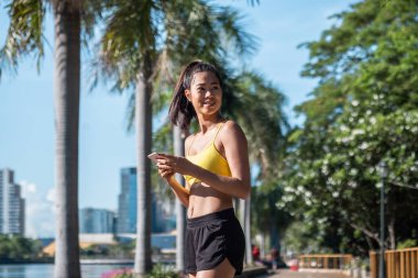 Gülümseyen sporcu kadın, arka planda nehir ve şehir binaları ile parkta yapılan antremandan sonra performansı izlemek için cep telefonu internet uygulamasına bakıyor.