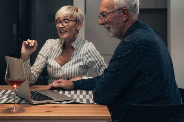 Evde dizüstü bilgisayar kullanan mutlu bir çift. Gülümseyen bir çift masada oturuyor, video konuşmaları yapıyorlar dizüstü bilgisayarlarını kullanıyorlar ve evde şarap içiyorlar. Karantina, Coronavirus, COVID-19, tecrit, evde kal.