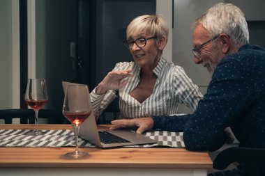 Evde dizüstü bilgisayar kullanan mutlu bir çift. Gülümseyen bir çift masada oturuyor, video konuşmaları yapıyorlar dizüstü bilgisayarlarını kullanıyorlar ve evde şarap içiyorlar. Karantina, Coronavirus, COVID-19, tecrit, evde kal.
