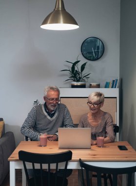 Mutlu son sınıf çifti evde dizüstü bilgisayar kullanıyor ve smoothie içiyor. Gülümseyen çift konuşuyor ve mutfak masasında oturuyor.