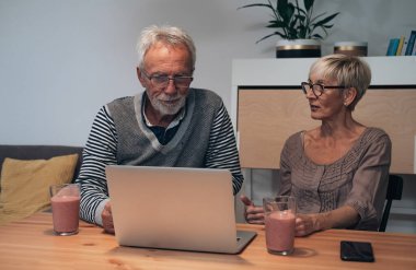 Üst düzey çift evde dizüstü bilgisayar kullanıyor ve smoothie içiyor. Ciddi bir çift mutfak masasında oturup konuşuyor.