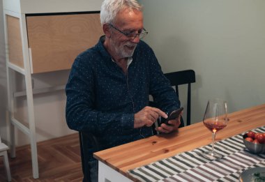 Evde telefon kullanan kıdemli bir adam. Yakışıklı, gülümseyen olgun bir adam yemek masasında oturuyor, şarap içiyor ve mutfakta akıllı telefon kullanıyor.