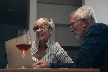 Evde dizüstü bilgisayar kullanan mutlu bir çift. Gülümseyen olgun bir çift masada oturuyor, bilgisayar kullanarak video görüşmesi yapıyor ve evde şarap içiyor.