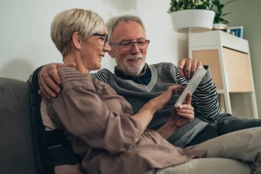 Evde dijital tablet kullanan yaşlı çifti kucaklamak. Oturma odasındaki kanepede oturan mutlu çift konuşuyor, gülümsüyor, beyaz taşınabilir bilgisayarda görünüp bakıyor.