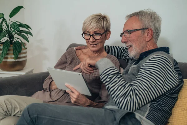 Embracing senior couple using digital tablet together at home. Happy senior couple sitting at couch at living room, talking, smiling, showing and looking in white portable computer