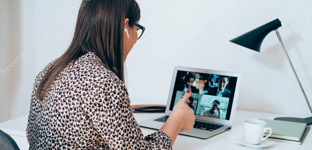 Businesswoman talking on video call and showing thumb up to laptop on the online meeting with colleagues. Unrecognizable business woman discuss with clients or coworkers at web conference using a laptop and bluetooth earphones while working at home