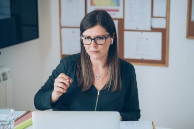 Gözlüklü ve bluetooth kulaklıklı öğretmen masada oturuyor ve dizüstü bilgisayarına bakıyor, öğrencileri kalemle işaret ediyor. Coronavirüs salgını sırasında e-öğrenim öğrencileri için online ders veren kadın profesör.